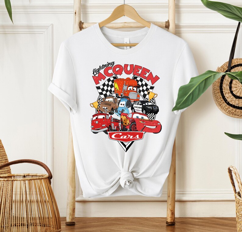 May include: White t-shirt featuring a colorful graphic of characters from the movie Cars, including Lightning McQueen. The design includes the text "Lightning McQueen" and "Cars" in red and black. The shirt is hanging on a wooden hanger.