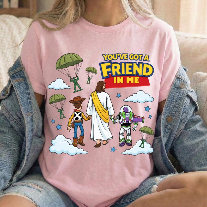 Peut inclure: Un t-shirt rose clair avec un motif repr&eacute;sentant une silhouette en robe blanche tenant la main d'une poup&eacute;e cow-boy et d'une poup&eacute;e ranger de l'espace. Le design inclut des soldats verts avec des parachutes, des nuages blancs, des &eacute;toiles bleues et le texte YOU'VE GOT A FRIEND IN ME.