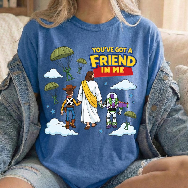 Peut inclure: Un t-shirt bleu avec une impression graphique repr&eacute;sentant une figure centrale en robe blanche avec une &eacute;charpe jaune, tenant la main d'une poup&eacute;e cow-boy et d'un jouet ranger de l'espace. Des parachutistes descendent avec le texte YOU'VE GOT A FRIEND IN ME.