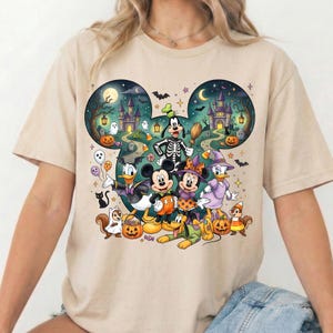 K&ouml;nnte beinhalten: Ein beiges Kurzarm-T-Shirt mit einem gro&szlig;en Grafikdruck von Mickey Maus, Minnie Maus, Donald Duck, Daisy Duck, Goofy sowie Chip und Chap in Halloween-Kost&uuml;men. Das Motiv ist in einer Mickey-f&ouml;rmigen Umrandung mit einem Gruselschloss im Hintergrund.