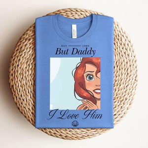 May include: A light blue t-shirt with a graphic of Ariel from The Little Mermaid. The shirt features the text "Est. 1989 But Daddy I Love Him" in black script. A seashell graphic is at the bottom.