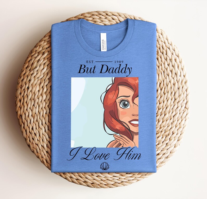 May include: A light blue t-shirt with a graphic of Ariel from The Little Mermaid. The shirt features the text "Est. 1989 But Daddy I Love Him" in black script. A seashell graphic is at the bottom.
