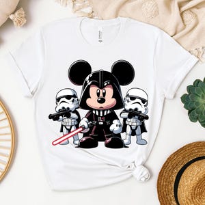 K&ouml;nnte beinhalten: Wei&szlig;es T-Shirt mit einer Cartoon-Illustration von Mickey Mouse als Darth Vader, flankiert von zwei Sturmtruppen. Mickey h&auml;lt ein rotes Lichtschwert. Das Design ist eine spielerische Mischung aus Disney und Star Wars.