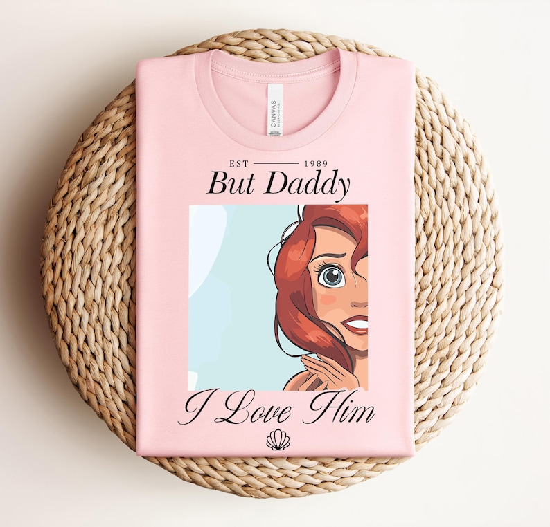 May include: A pale pink t-shirt featuring the text "But Daddy I Love Him" and a cartoon illustration of a character with red hair and blue eyes. The shirt is folded on a woven mat.