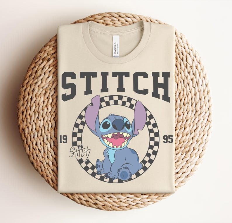 May include: Beige t-shirt featuring a graphic of Stitch from Lilo & Stitch. The design includes the word "STITCH" in dark gray block letters, the year "1995", and a checkered circle around Stitch. The shirt is folded on a woven surface.