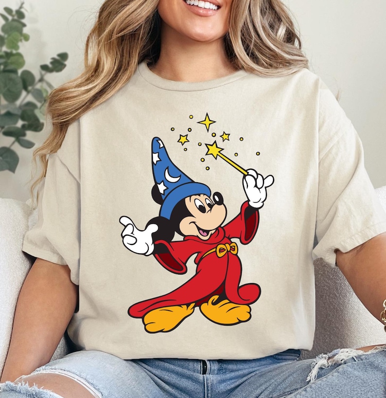May include: Beige t-shirt featuring a cartoon illustration of Mickey Mouse dressed as a wizard. Mickey is wearing a blue hat with white stars, a red robe, and yellow shoes. He is holding a wand with yellow stars emanating from it.