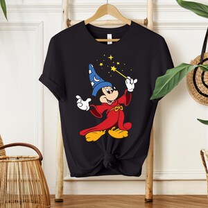 May include: Black t-shirt featuring a cartoon illustration of Mickey Mouse dressed as a wizard. Mickey is wearing a blue wizard hat with white stars and a red robe, holding a wand with yellow stars. The shirt is hanging on a wooden hanger.