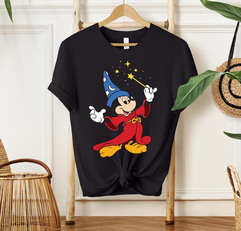 May include: Black t-shirt featuring a cartoon illustration of Mickey Mouse dressed as a wizard. Mickey is wearing a blue wizard hat with white stars and a red robe, holding a wand with yellow stars. The shirt is hanging on a wooden hanger.