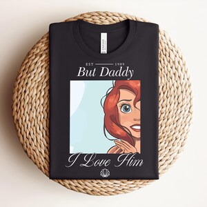 May include: Black t-shirt with a cartoon illustration of a red-haired character and the text "But Daddy I Love Him" and the year "1989". A seashell graphic is included. The shirt is folded on a woven surface.