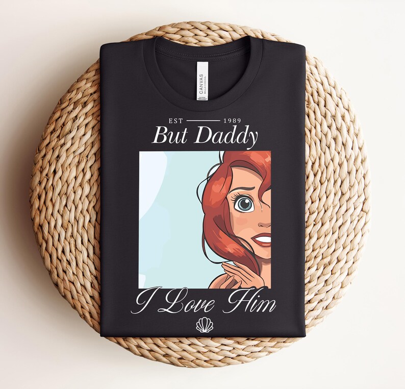 May include: Black t-shirt with a cartoon illustration of a red-haired character and the text "But Daddy I Love Him" and the year "1989". A seashell graphic is included. The shirt is folded on a woven surface.