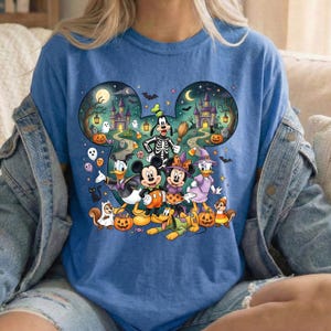 K&ouml;nnte beinhalten: Ein blaues T-Shirt mit einem gro&szlig;en, Mickey-Maus-ohrenf&ouml;rmigen Aufdruck, der eine Halloween-Szene zeigt. Das Design enth&auml;lt Zeichentrickfiguren in Kost&uuml;men, K&uuml;rbisse, ein Spukschloss, Flederm&auml;use und einen Mond vor einem dunklen Nachthimmel.