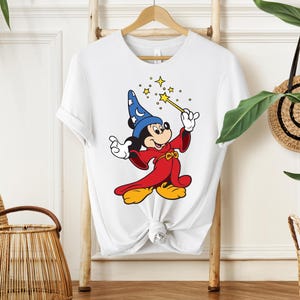 May include: White t-shirt featuring a cartoon illustration of Mickey Mouse dressed as a wizard. Mickey is wearing a blue hat with stars, a red robe, and yellow shoes, holding a wand with sparkling stars.