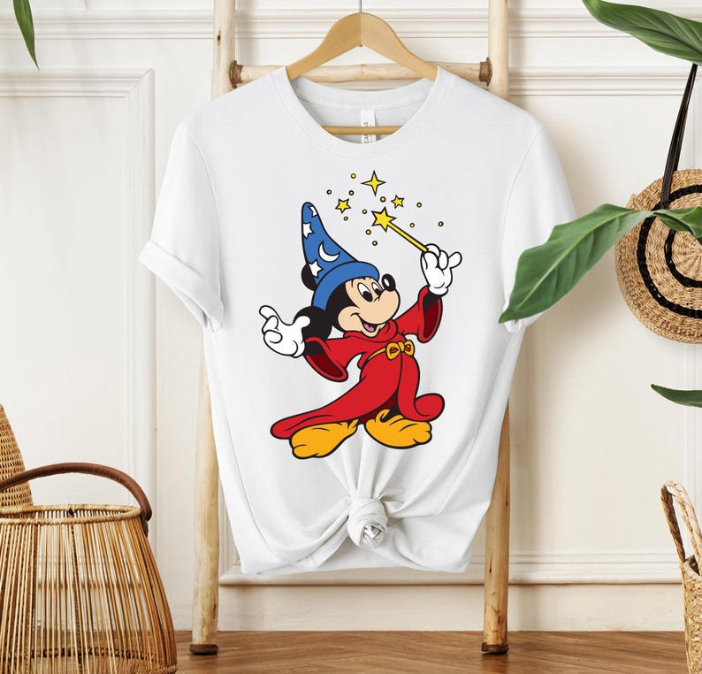 May include: White t-shirt featuring a cartoon illustration of Mickey Mouse dressed as a wizard. Mickey is wearing a blue hat with stars, a red robe, and yellow shoes, holding a wand with sparkling stars.