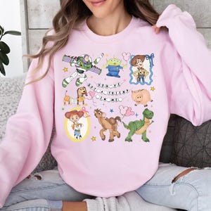 May include: A light pink sweatshirt featuring colorful cartoon characters from the movie Toy Story. The design includes Buzz Lightyear, Woody, Jessie, and other characters, with the text "You've Got a Friend in Me".