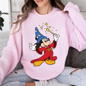 May include: Pink sweatshirt featuring a cartoon illustration of Mickey Mouse dressed as a wizard. Mickey is wearing a blue hat with white stars, a red robe, and holding a wand with a yellow star. The sweatshirt is a solid light pink color.