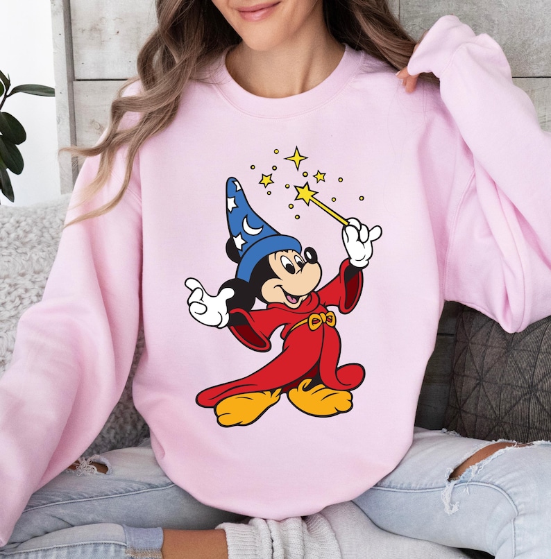 May include: Pink sweatshirt featuring a cartoon illustration of Mickey Mouse dressed as a wizard. Mickey is wearing a blue hat with white stars, a red robe, and holding a wand with a yellow star. The sweatshirt is a solid light pink color.