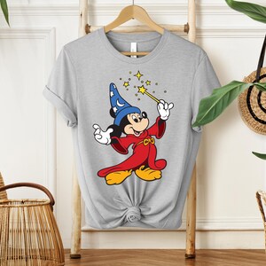 May include: A light gray t-shirt featuring a cartoon image of Mickey Mouse dressed as a wizard. Mickey is wearing a blue hat with white stars, a red robe, and yellow shoes, holding a gold wand with stars.