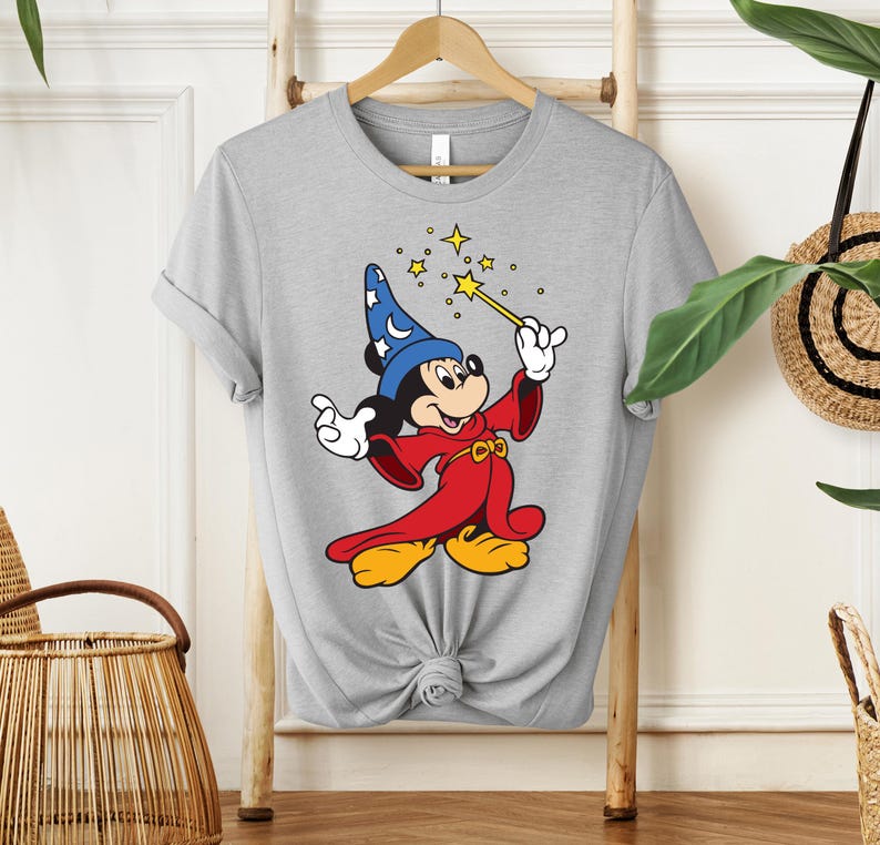 May include: A light gray t-shirt featuring a cartoon image of Mickey Mouse dressed as a wizard. Mickey is wearing a blue hat with white stars, a red robe, and yellow shoes, holding a gold wand with stars.