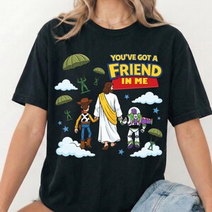 Peut inclure: Un t-shirt noir avec une impression graphique repr&eacute;sentant une figure centrale en robe blanche avec une &eacute;charpe jaune, tenant la main d'une poup&eacute;e cow-boy et d'une poup&eacute;e ranger de l'espace. Au-dessus, le texte YOU'VE GOT A FRIEND IN ME, avec des soldats jouets verts descendant en parachute et des nuages blancs.