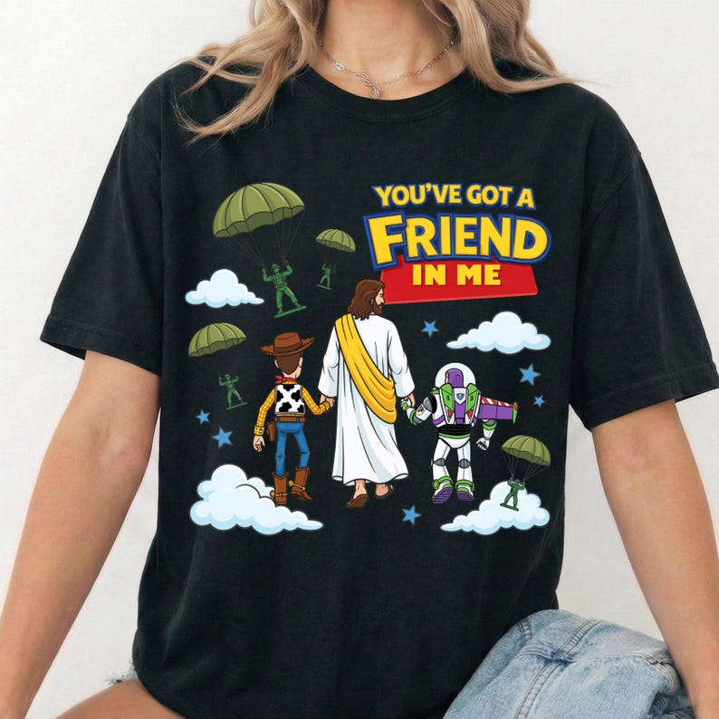 Peut inclure: Un t-shirt noir avec une impression graphique repr&eacute;sentant une figure centrale en robe blanche avec une &eacute;charpe jaune, tenant la main d'une poup&eacute;e cow-boy et d'une poup&eacute;e ranger de l'espace. Au-dessus, le texte YOU'VE GOT A FRIEND IN ME, avec des soldats jouets verts descendant en parachute et des nuages blancs.