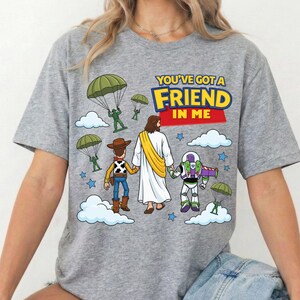 Peut inclure: Un t-shirt gris chin&eacute; avec une illustration repr&eacute;sentant une poup&eacute;e cow-boy, une poup&eacute;e ranger de l'espace et une silhouette en robe blanche avec une &eacute;charpe jaune marchant ensemble. Le texte indique YOU'VE GOT A FRIEND IN ME, avec des parachutistes verts.