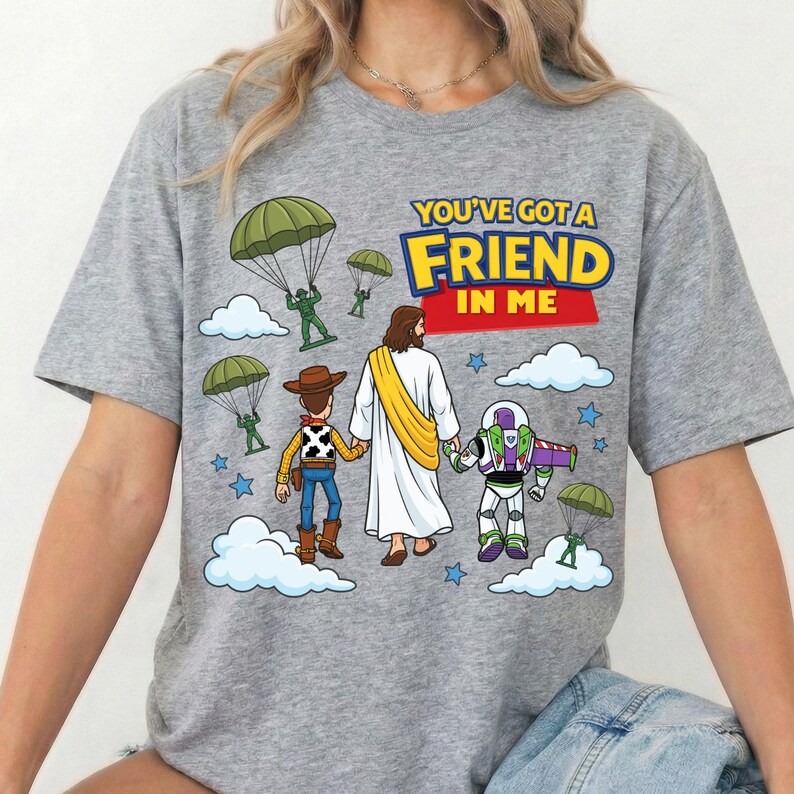 Peut inclure: Un t-shirt gris chin&eacute; avec une illustration repr&eacute;sentant une poup&eacute;e cow-boy, une poup&eacute;e ranger de l'espace et une silhouette en robe blanche avec une &eacute;charpe jaune marchant ensemble. Le texte indique YOU'VE GOT A FRIEND IN ME, avec des parachutistes verts.