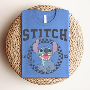 May include: Blue t-shirt featuring a graphic of Stitch from Lilo & Stitch. The design includes the word "STITCH" in bold, gray letters above a circular image of Stitch. The image is surrounded by a checkered pattern, with the numbers "19" and "95" on either side.
