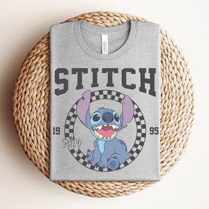 May include: A gray t-shirt featuring the word "STITCH" in dark gray block letters above a cartoon image of Stitch inside a black and white checkered circle. The numbers "19" and "95" flank the image.