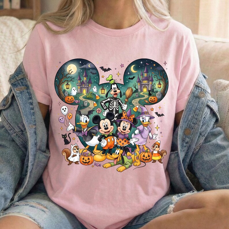 K&ouml;nnte beinhalten: Ein hellrosa T-Shirt mit einer Grafik von Disney-Figuren in Halloween-Kost&uuml;men, darunter Mickey, Minnie, Donald, Goofy und Pluto. Das Design ist wie Mickey-Ohren geformt und zeigt ein Spukschloss, K&uuml;rbisse, Geister und Flederm&auml;use.