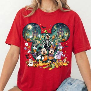 K&ouml;nnte beinhalten: Ein rotes T-Shirt mit einer gro&szlig;en Grafik in Form von Mickey-Mouse-Ohren, die eine Halloween-Szene zeigt. Das Design enth&auml;lt Zeichentrickfiguren in Kost&uuml;men, ein Spukschloss, K&uuml;rbisse, Geister, Flederm&auml;use und einen Mond vor einem dunklen Nachthimmel.