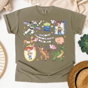 May include: Olive green t-shirt featuring colorful cartoon characters from a popular animated movie. The shirt includes Buzz Lightyear, an alien, Woody, Jessie, Hamm, Bullseye, Rex, and the phrase "You've Got a Friend in Me."