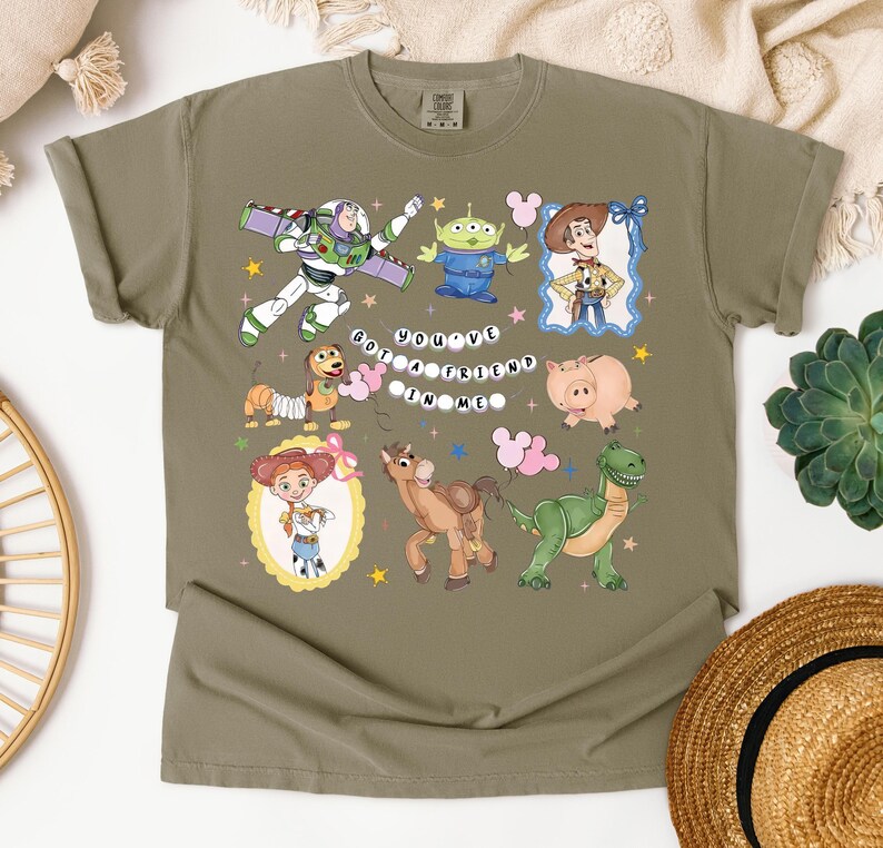 May include: Olive green t-shirt featuring colorful cartoon characters from a popular animated movie. The shirt includes Buzz Lightyear, an alien, Woody, Jessie, Hamm, Bullseye, Rex, and the phrase "You've Got a Friend in Me."
