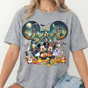 K&ouml;nnte beinhalten: Ein graues T-Shirt mit einem gro&szlig;en, Mickey-Maus-f&ouml;rmigen Aufdruck, der eine Halloween-Szene zeigt. Das Motiv enth&auml;lt Zeichentrickfiguren in Kost&uuml;men, K&uuml;rbisse, Geister, Flederm&auml;use und ein Schloss vor einem dunklen Nachthimmel.