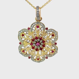 May include: A gold-toned pendant necklace featuring a floral design. The pendant is adorned with red gemstones and clear crystals, set within an intricate filigree pattern. The necklace has a delicate gold chain.
