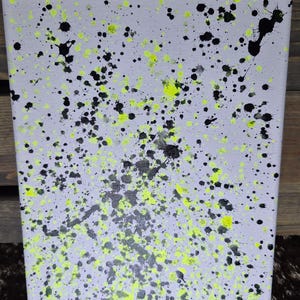 May include: Abstract art canvas with a light gray background, splattered with black and neon yellow paint. The artwork features a textured surface and a unique, modern design. Signed "Sierra".