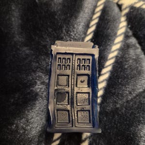 May include: A miniature blue and grey TARDIS model, a fictional time machine and spacecraft from the British science fiction television programme Doctor Who. The model has detailed door panels and windows.