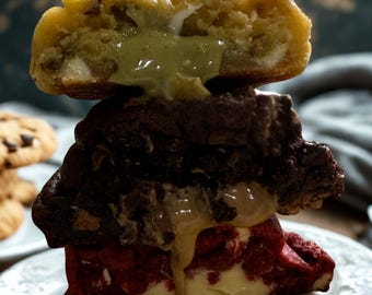 May include: A stack of three gourmet cookies on a white plate. The top cookie is golden with a green filling, the middle is dark chocolate, and the bottom is red velvet with a creamy filling. Chocolate chip cookies are in the background.