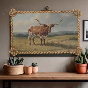 May include: A framed canvas print featuring a longhorn steer in a grassy field under a cloudy sky. The artwork is bordered by a rope-like frame. The print is displayed above a wooden surface with potted plants.