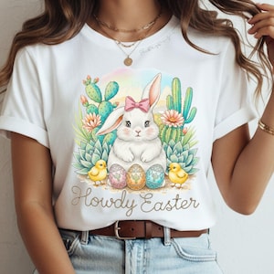 May include: White t-shirt featuring a watercolor design with a bunny wearing a pink bow, surrounded by cacti, flowers, and Easter eggs. Two yellow chicks flank the eggs. The text "Howdy Easter" is below the design.