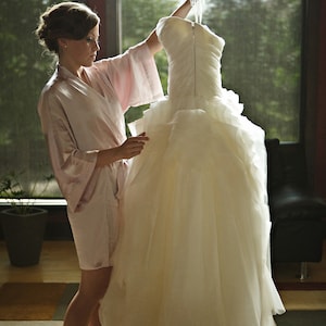 May include: A woman in a pink robe is holding a white wedding dress on a hanger. The dress is very full and has a lot of layers. The woman is standing in front of a window with a view of trees.