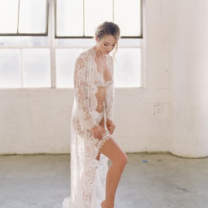 May include: A white lace robe with long sleeves and a delicate floral pattern. The robe is open in the front, revealing a white lace bra and matching underwear. The model is standing in a white room with a large window in the background.