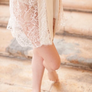 Op de afbeelding: Een witte kanten robe met een satijnen ceintuur geknoopt in de taille. De robe wordt gedragen over een witte short.