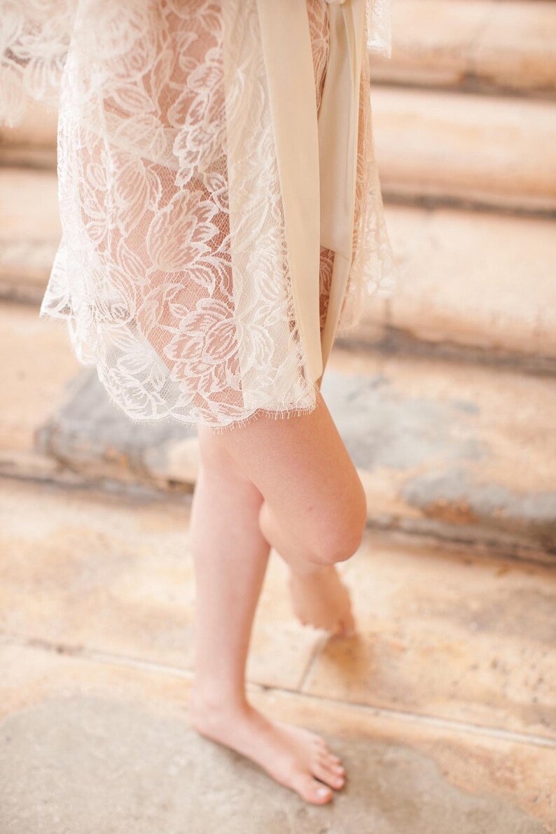 Op de afbeelding: Een witte kanten robe met een satijnen ceintuur geknoopt in de taille. De robe wordt gedragen over een witte short.