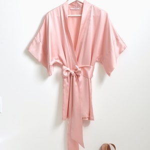 May include: A pale pink satin robe with a tie sash and short sleeves hangs on a white hanger against a white wall. Two pairs of nude ballet flats are on the floor below.