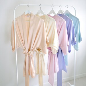 May include: Five satin robes in different pastel colors hanging on a white rack. The robes are in shades of peach, cream, pink, lavender, and light blue.