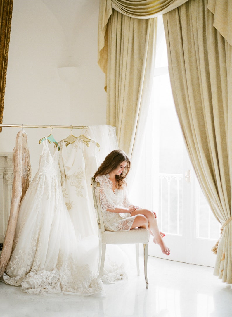 May include: A bride-to-be sits in a white lace robe in a room with large windows and white curtains. She is surrounded by several white wedding dresses hanging on a rack.