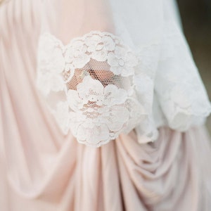 May include: A close-up of a white lace trim on a blush-colored fabric. The lace is delicate and intricate, with a floral pattern.