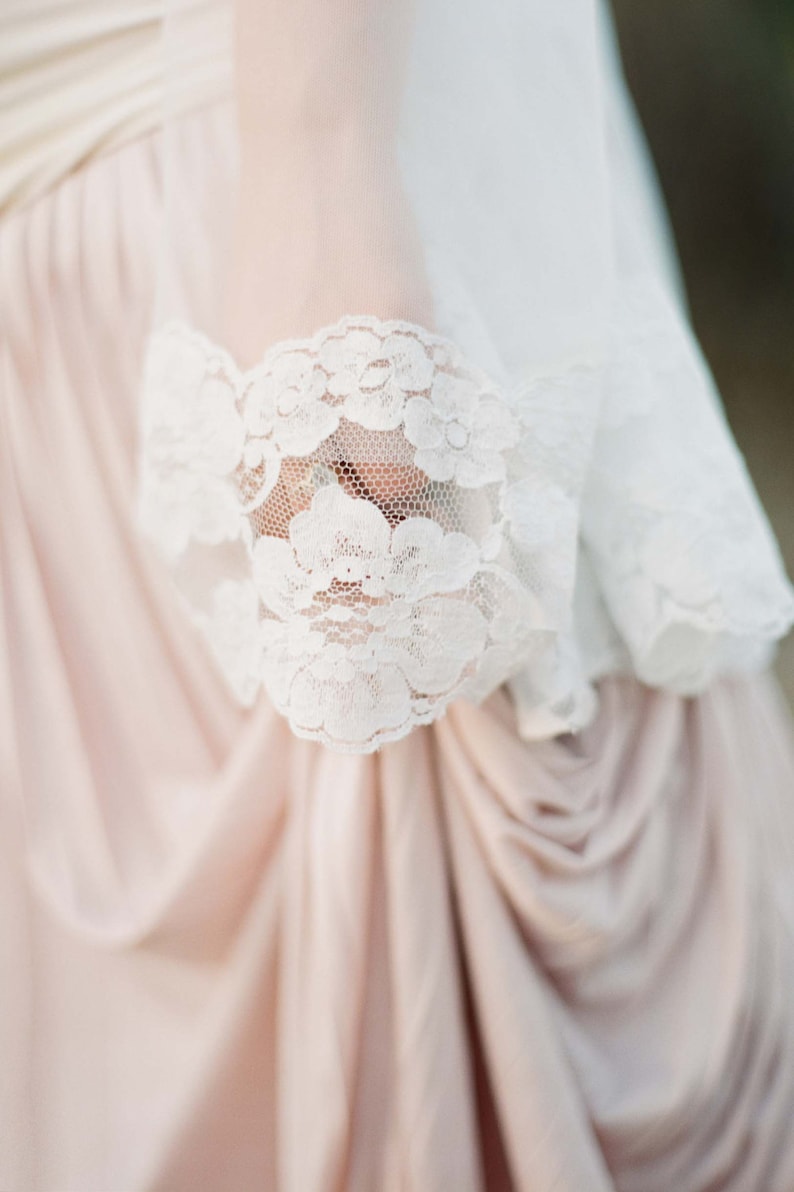 May include: A close-up of a white lace trim on a blush-colored fabric. The lace is delicate and intricate, with a floral pattern.