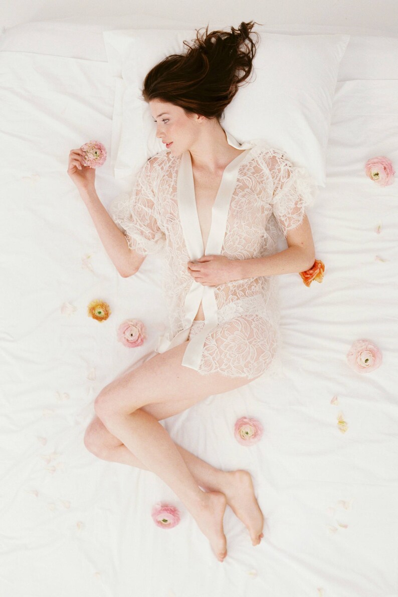 Op de afbeelding: Een vrouw in een witte kanten jurk ligt op een wit bed met roze bloemen die eromheen zijn verspreid.