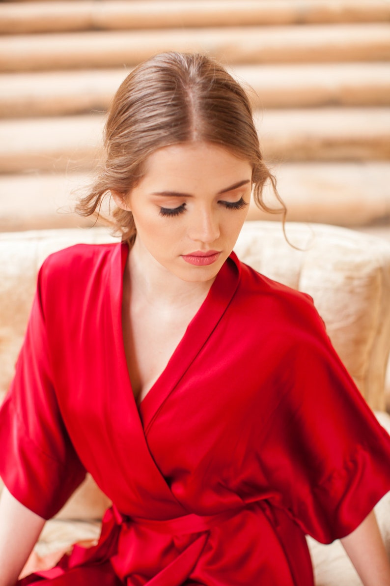 May include: A woman wearing a red satin robe with a deep V-neckline. The robe has long sleeves and a sash tie at the waist.